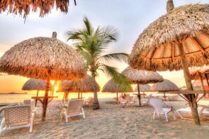 A beach with thatched umbrellas, lounge chairs, and a palm tree. - Hotel Santorini Kima Beach Resort (Santa Marta)