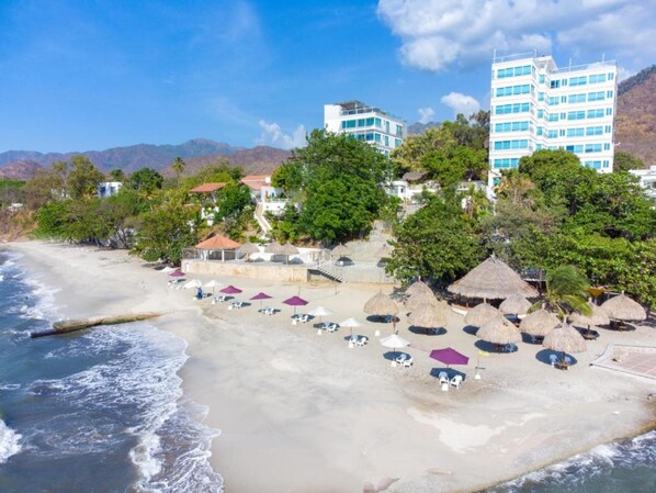 A beach with umbrellas and lounge chairs, a building with multiple floors, and a mountainous backdrop. - Hotel Santorini Kima Beach Resort (Santa Marta)