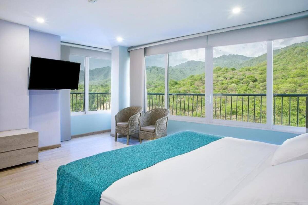 Une chambre d’hôtel avec un grand lit, une télévision, deux fauteuils en osier et un balcon offrant une vue sur la montagne.