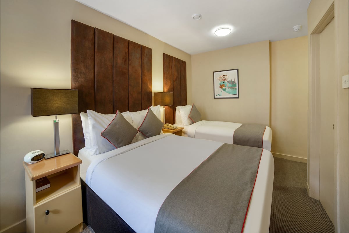 Habitación de hotel con dos camas, cabecera de madera, mesita de noche con lámpara, un cuadro enmarcado en la pared y suelo alfombrado.