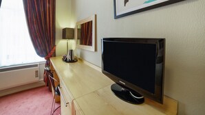 A hotel room with a wooden desk, a flat-screen TV, a lamp, a mirror, and a window with red curtains.