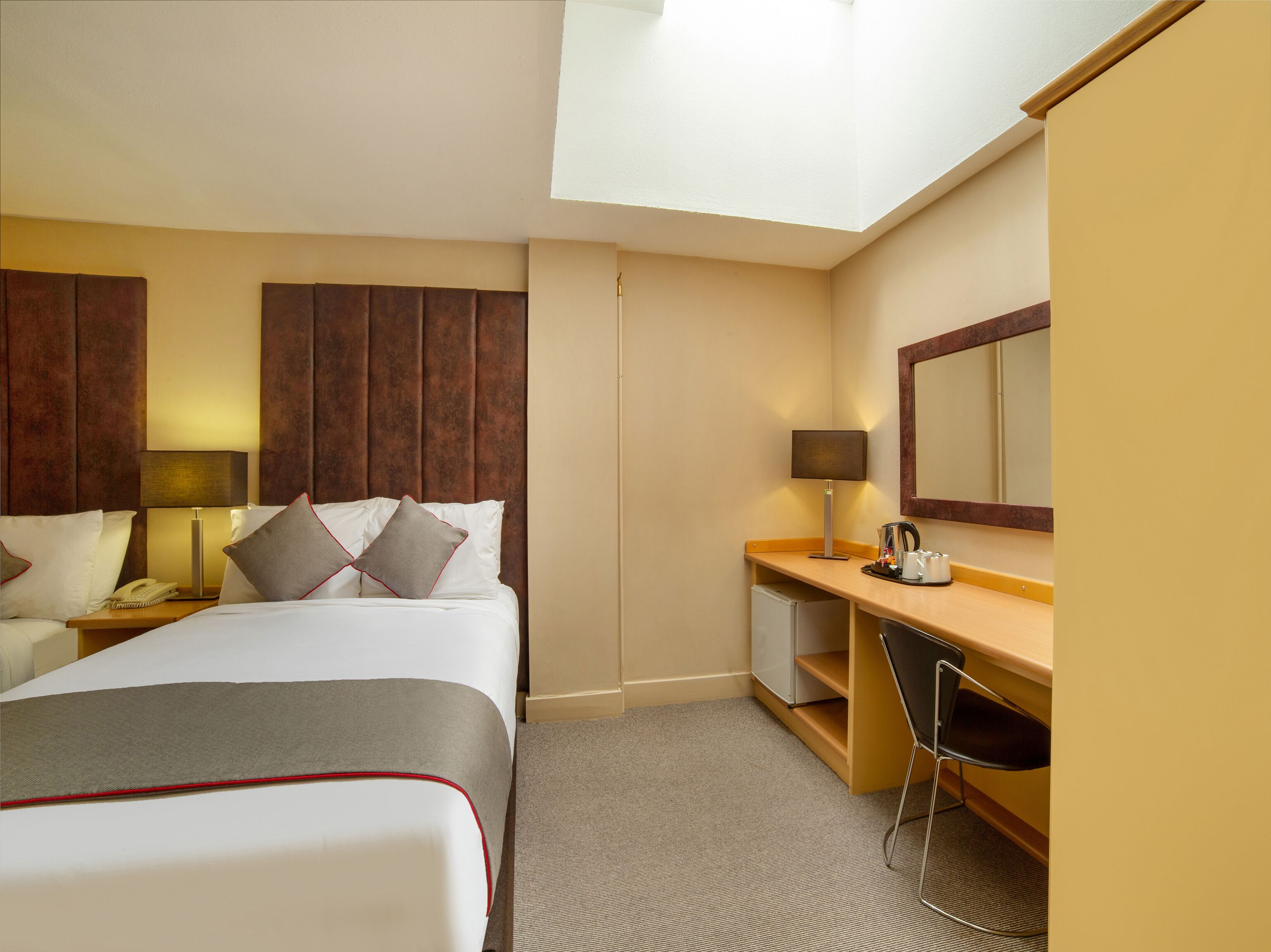 A hotel room with a bed, bedside table, lamp, desk, chair, and a mirror.