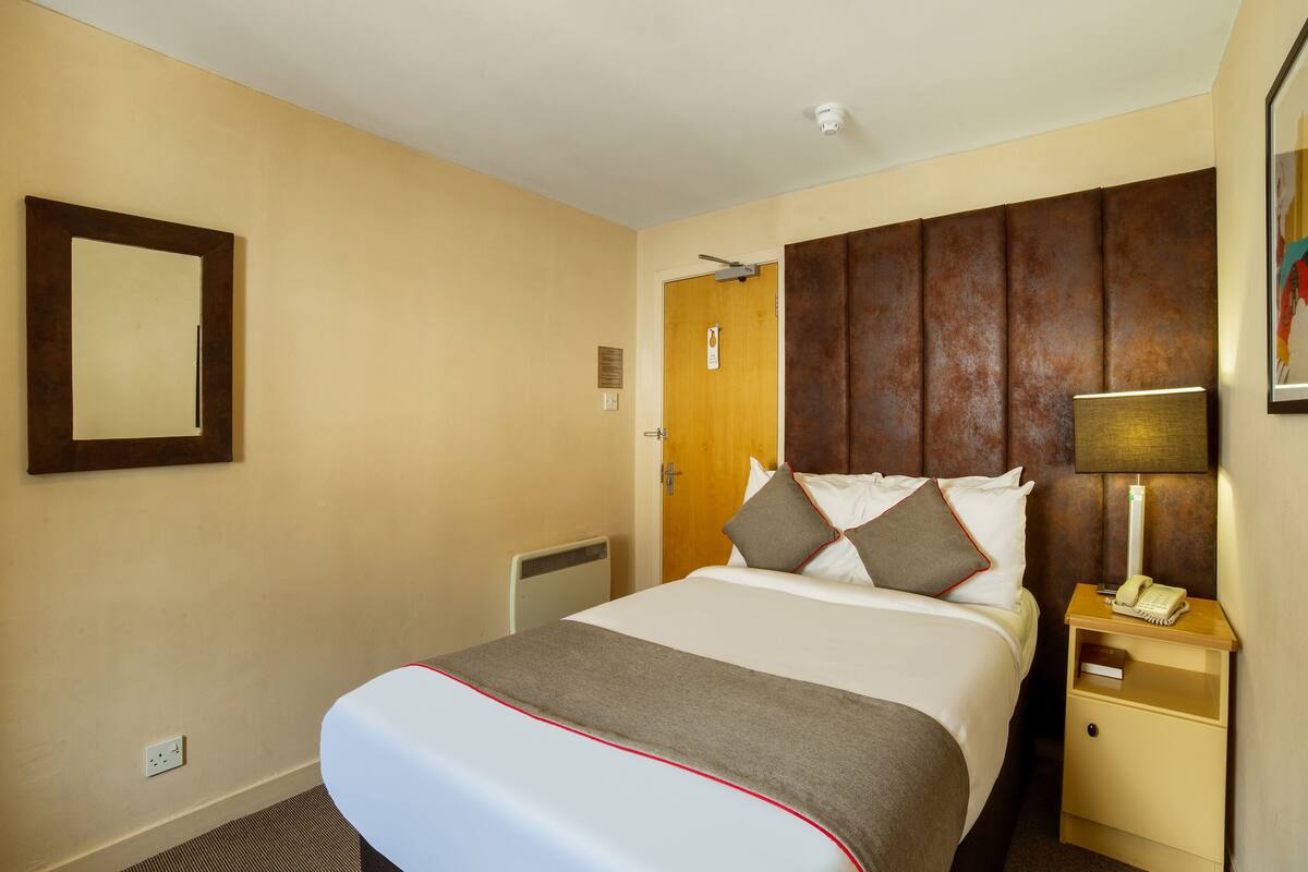 Habitación de hotel con cama, mesita de noche con lámpara, teléfono en la pared y una puerta.