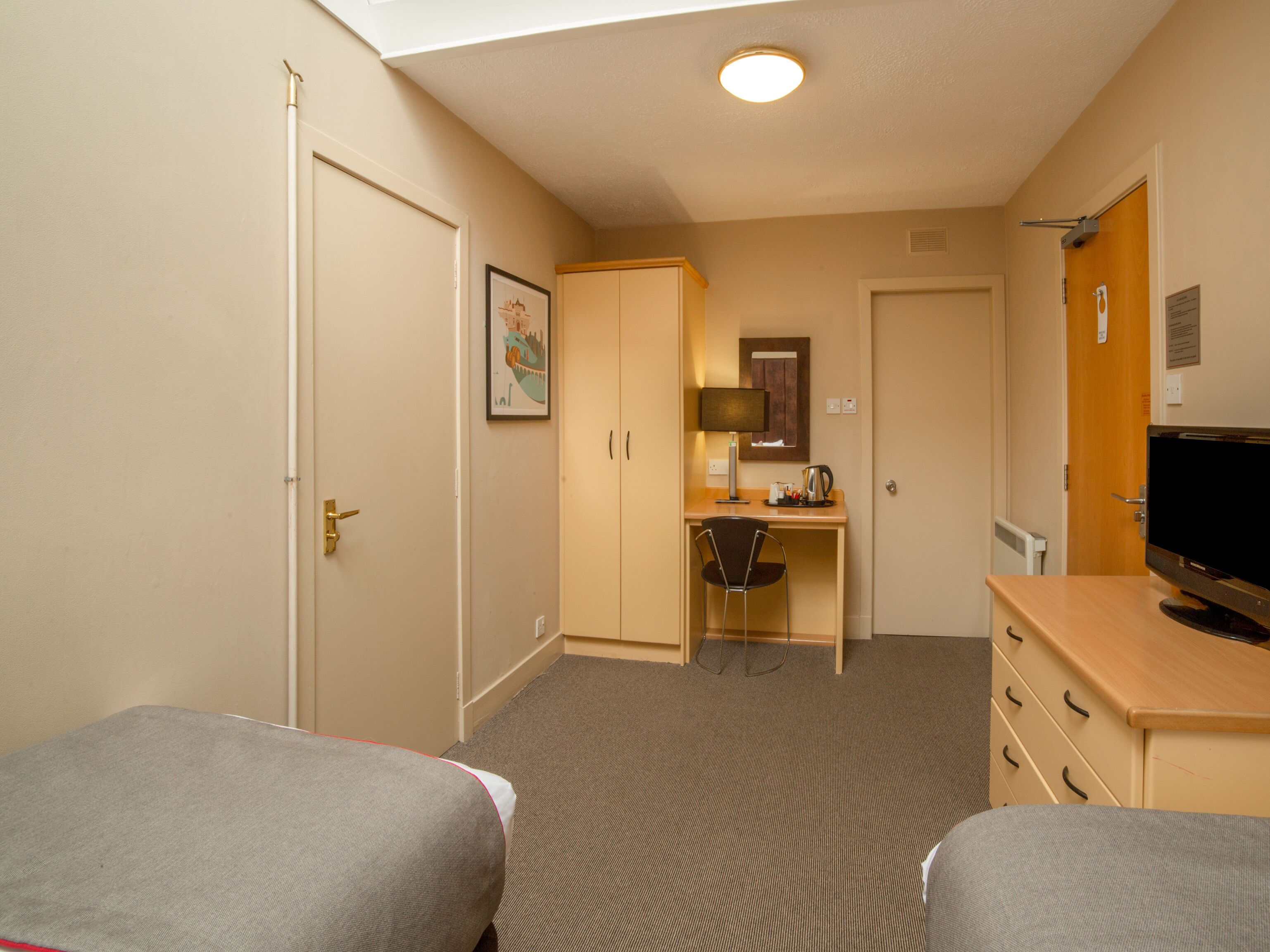 Una habitación de hotel con dos camas individuales, un escritorio con silla, un televisor, una kitchenette pequeña y una puerta que da a otra habitación.