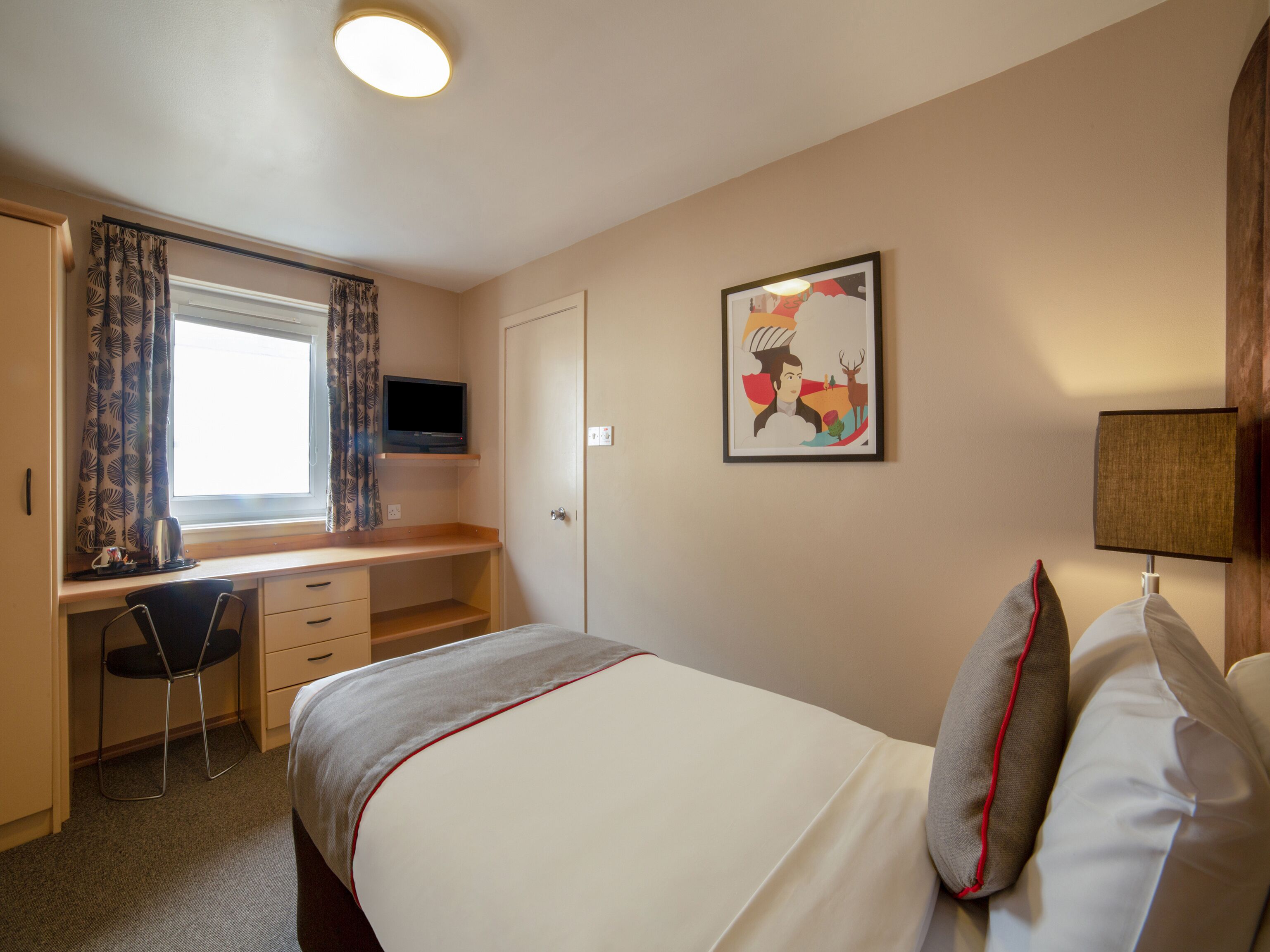 Hotelkamer met een bed, een bureau met stoel, een tv boven het bureau, een raam met gordijnen, een deur, een wandlamp en een ingelijst schilderij.