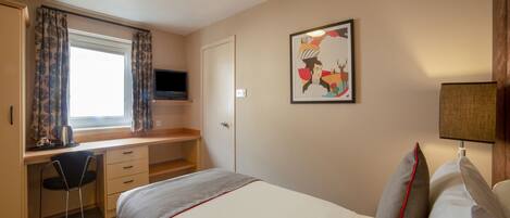 A hotel room with a bed, a desk with a chair, a TV mounted above the desk, a window with curtains, a door, a wall-mounted lamp, and a framed picture.