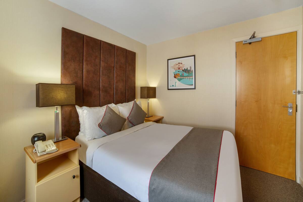 Habitación de hotel con una cama, dos mesitas de noche, una lámpara, un teléfono, una puerta y un cuadro colgado en la pared.