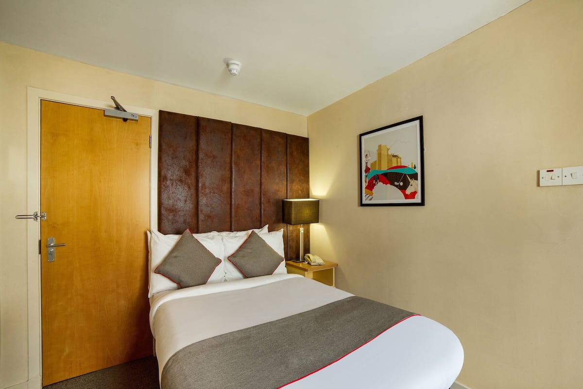 Una habitación de hotel con cama, puerta, mesita de noche con lámpara y una lámpara de pared.