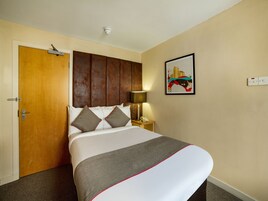 Una habitación de hotel con cama, puerta, mesita de noche con lámpara y una lámpara de pared.