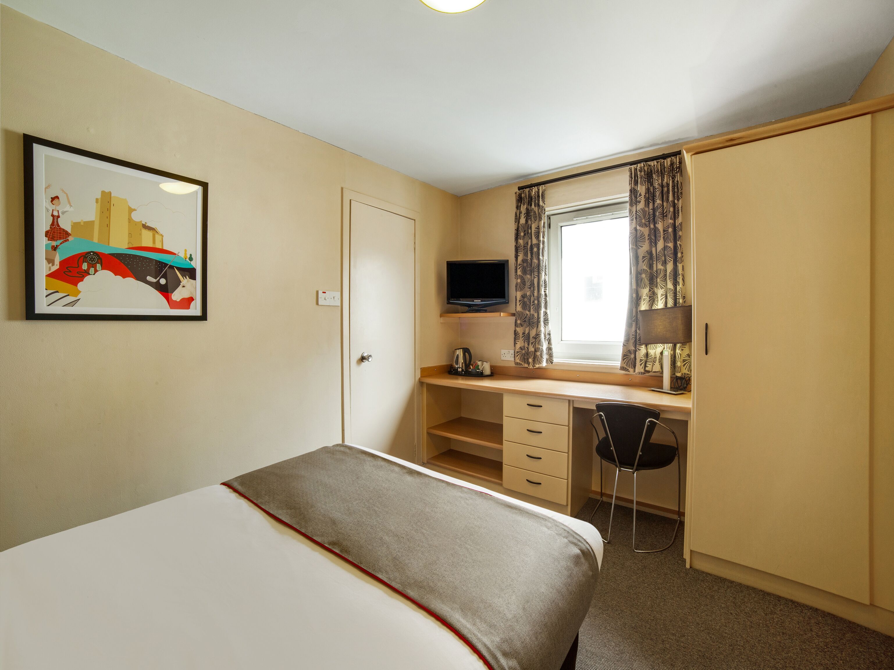 Een hotelkamer met een bed, een bureau met stoel, een televisie en een raam met gordijnen.