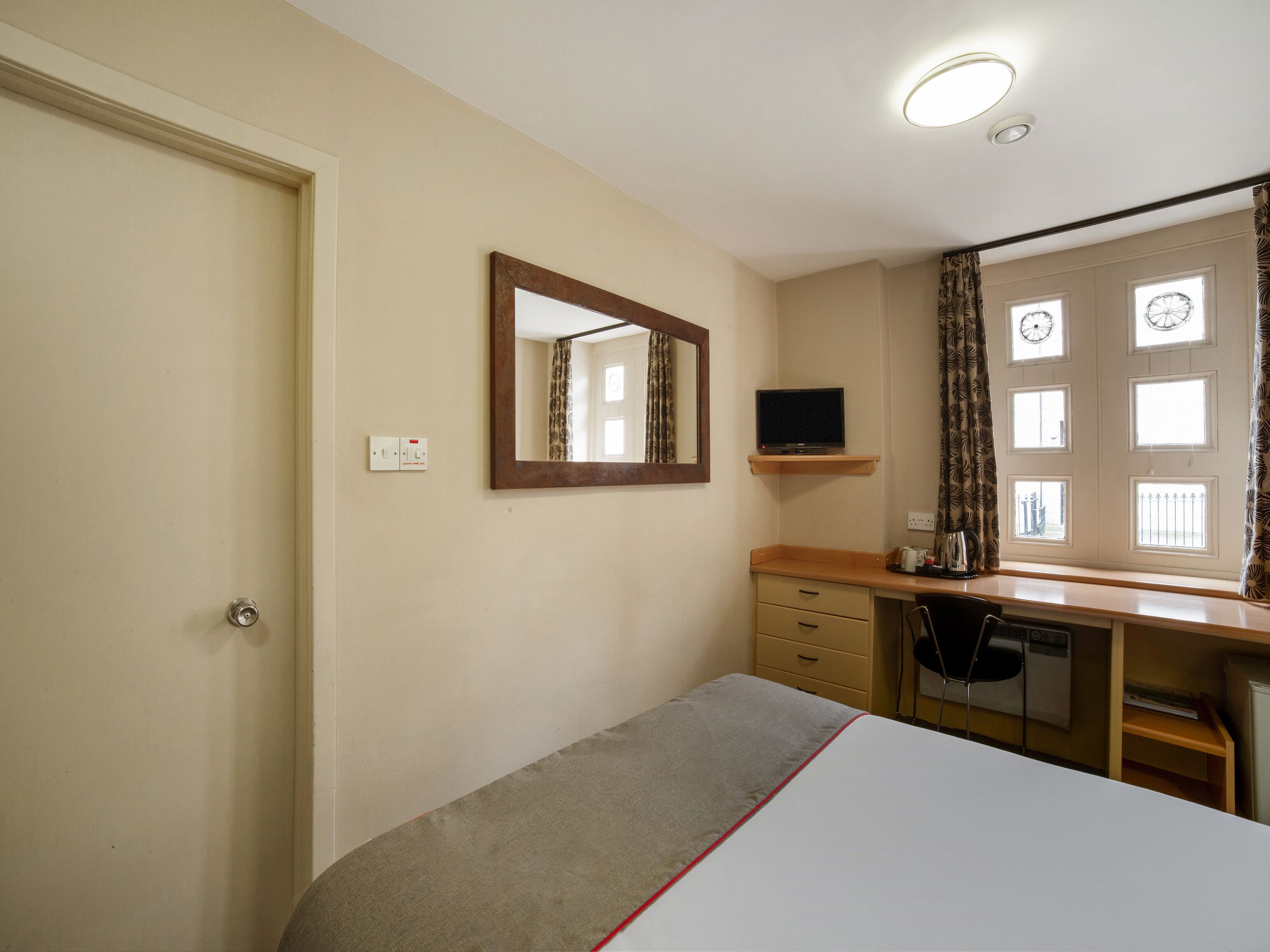 Ein Schlafzimmer mit einem Bett, einem Schreibtisch mit Stuhl, einem Spiegel, einem Fernseher und einem Fenster mit Vorhängen.