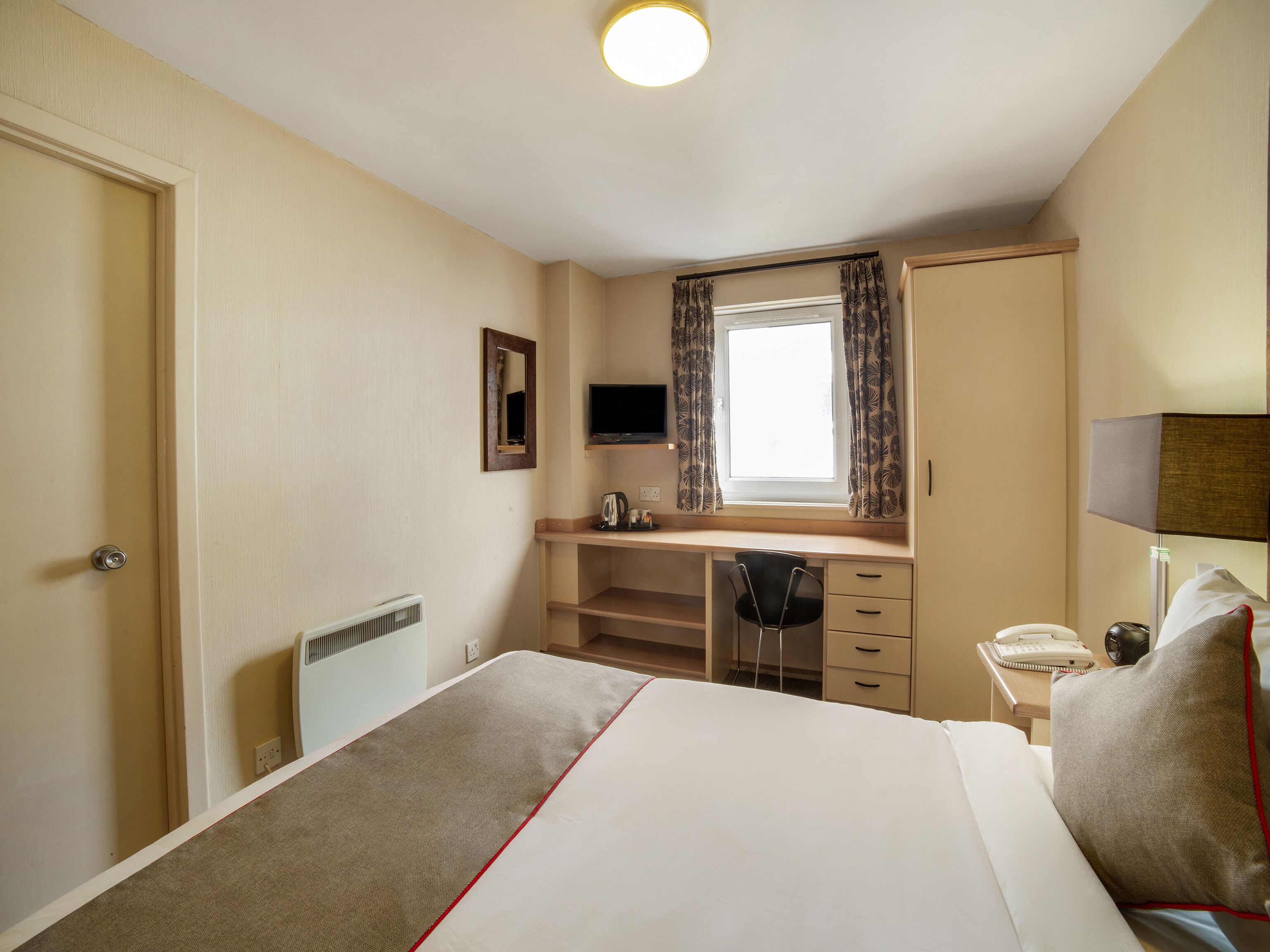 A hotel room with a bed, a desk, a chair, a TV, a window with curtains, and a bedside lamp.