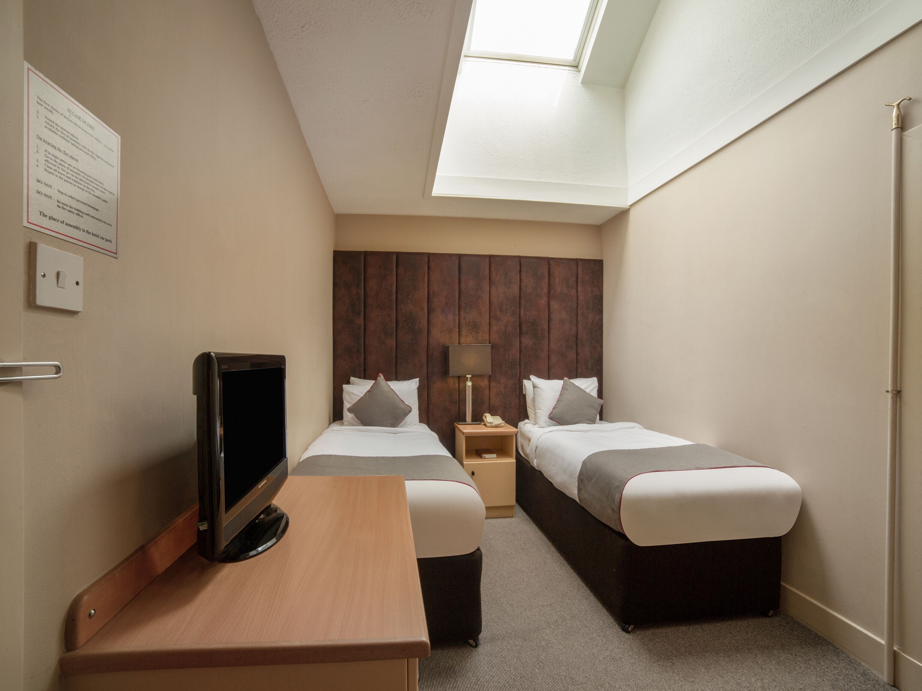A hotel room with two beds, a desk, a TV, a lamp, and a skylight.