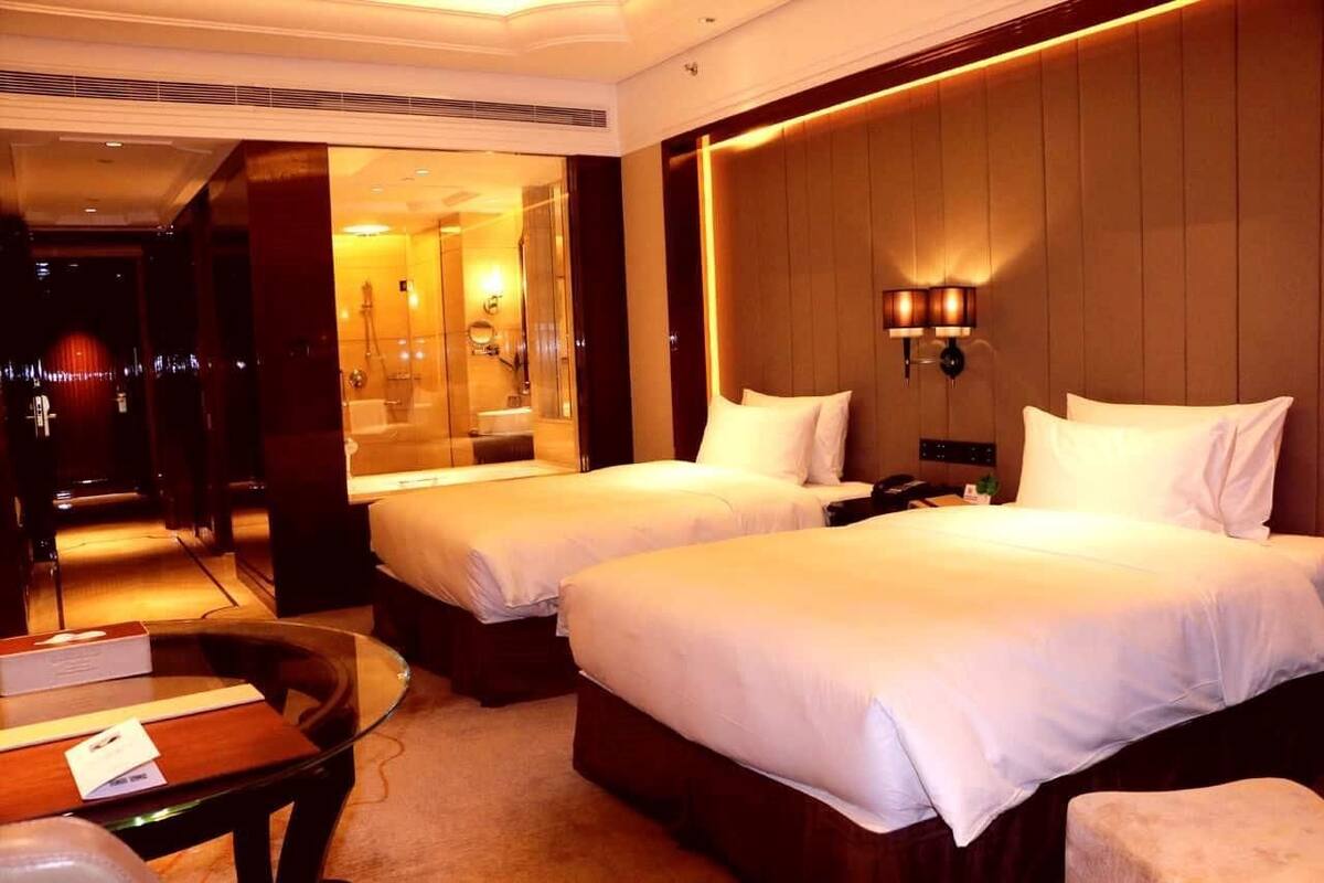 A hotel room with two beds, a sitting area with a table, and a bathroom visible through a glass door.