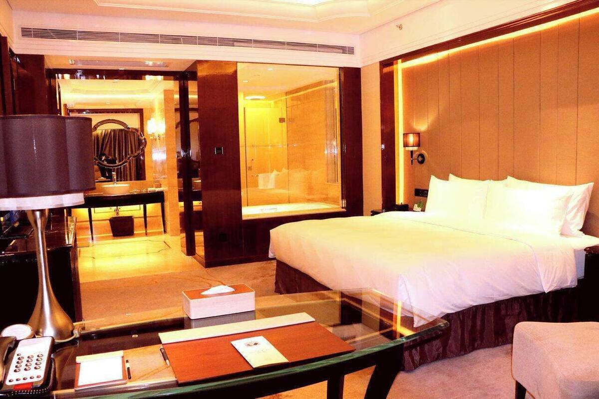 A hotel room with a large bed, a glass table with a phone and papers, a desk with a lamp, and a mirror.