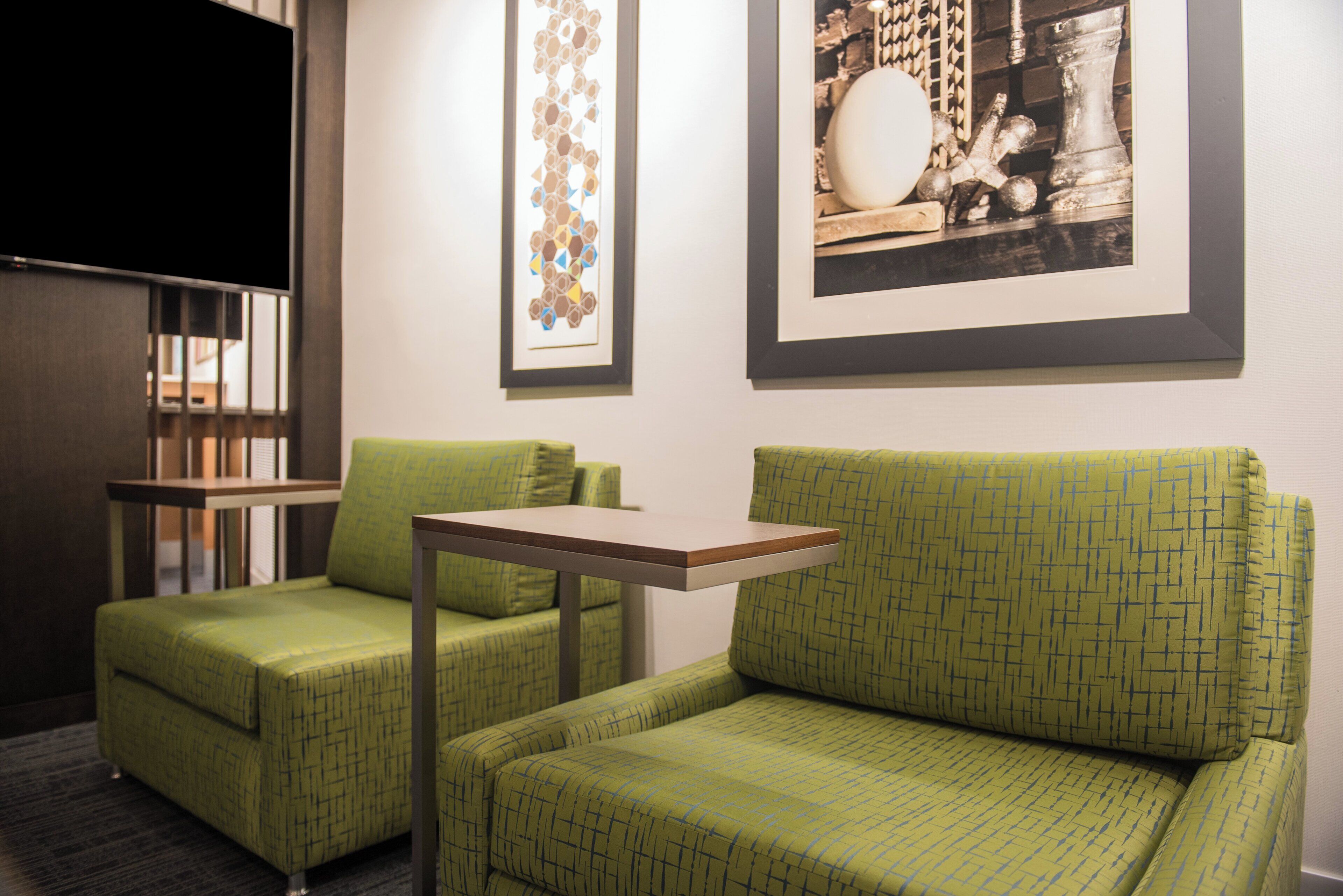 A room with two green upholstered chairs, a small table, and a wall-mounted flat-screen TV.