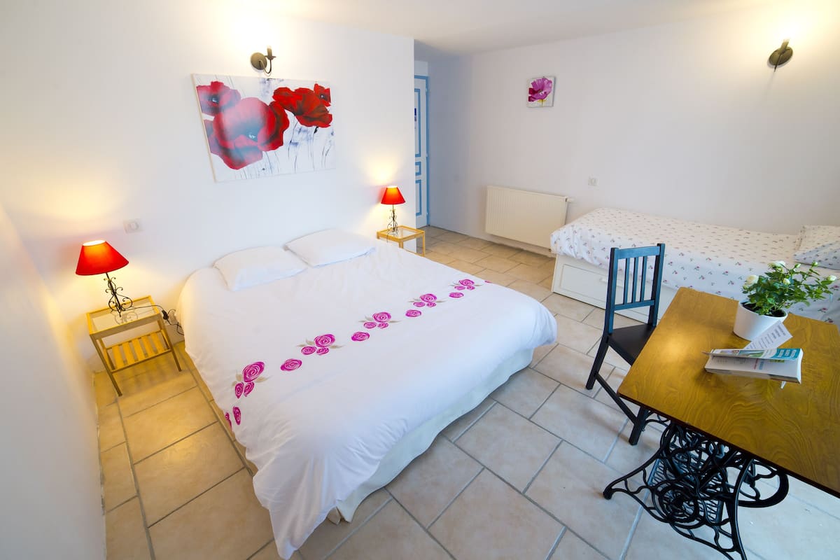 Una habitación de hotel con una cama, una mesa de madera con una silla, una mesita con una lámpara y una pared decorada con un cuadro floral.
