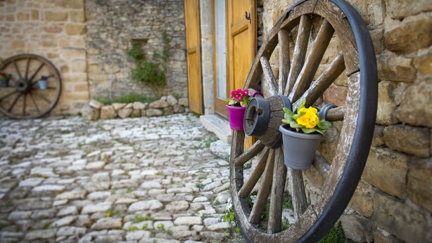 A rustic stone building with a large wooden wheel attached to the wall, two potted plants, and a cobblestone path.