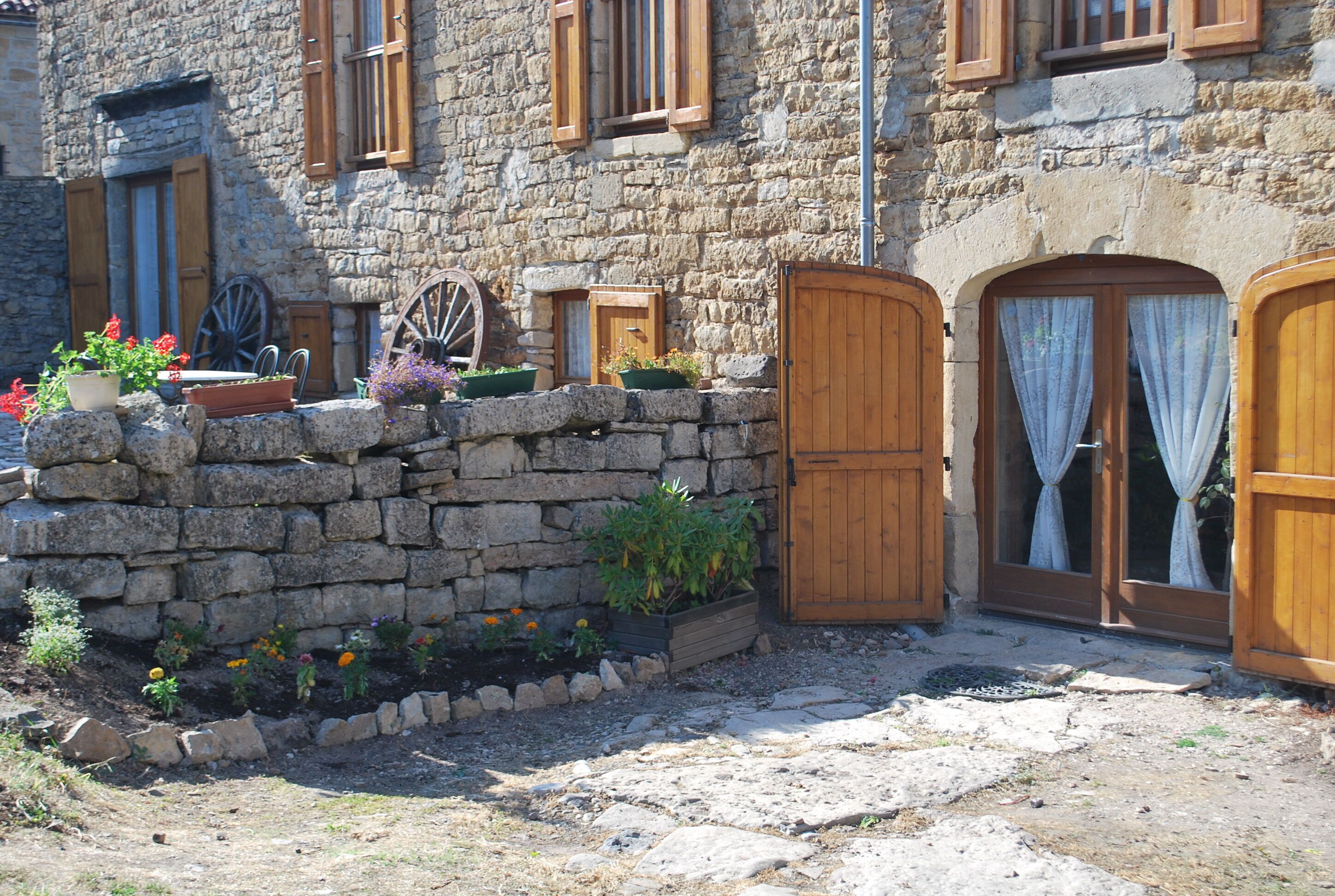 Un edificio de piedra con contraventanas de madera, una puerta de madera y un pequeño jardín con macetas y flores.