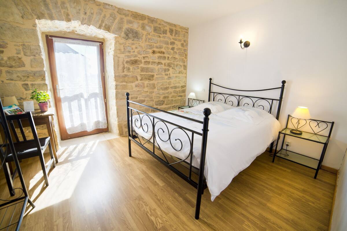 Un dormitorio con pared de piedra, cama con estructura metálica, piso de madera, escritorio con silla y una mesita con lámpara.