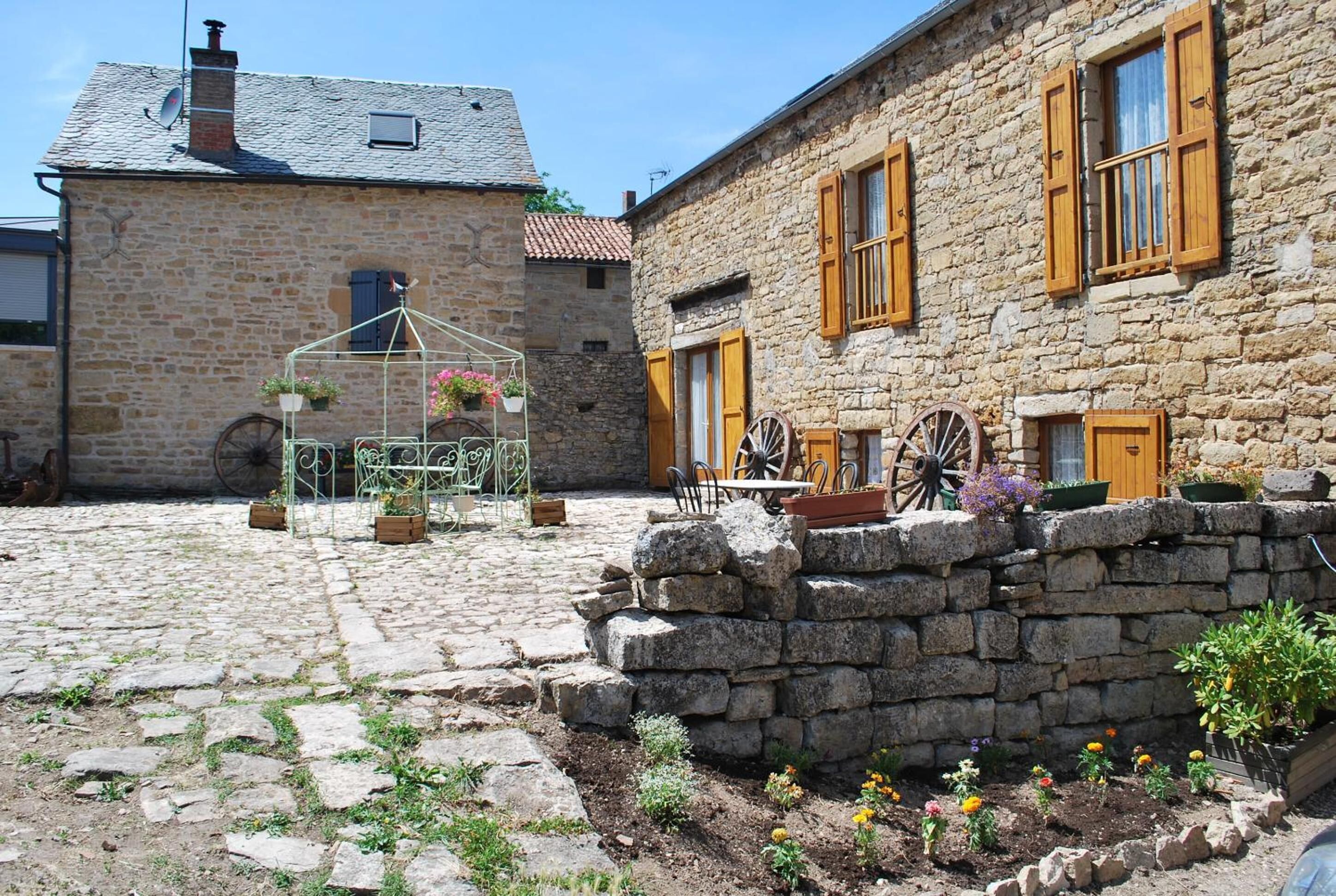Un patio de piedra con un muro de piedra rústico, una reja de metal y un pequeño jardín con flores y una carretilla.