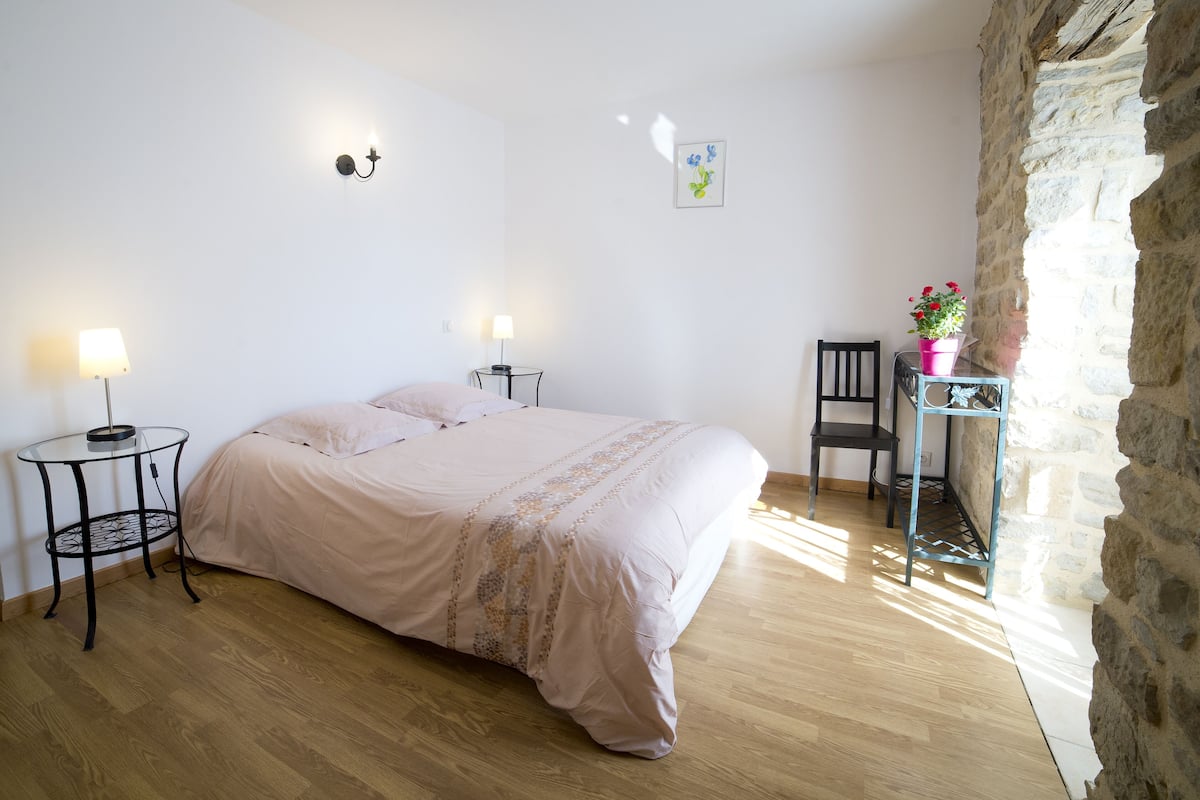 Un dormitorio con una cama, dos mesitas de noche con lámparas, una silla, una pequeña mesa con una maceta y una pared de piedra.