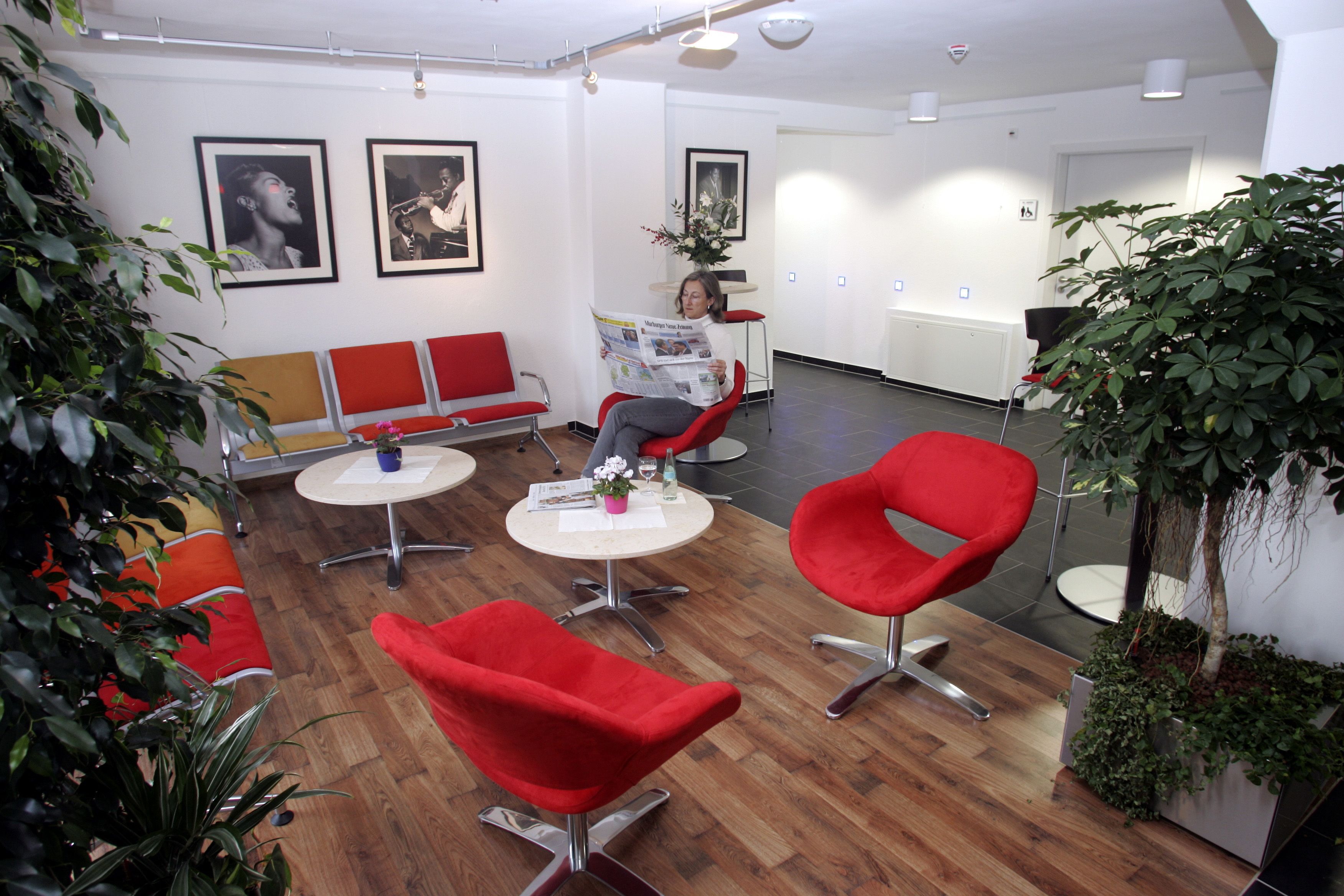 Een moderne wachtruimte met rode stoelen, een ronde tafel en een bank. Er hangen afbeeldingen in lijsten aan de muur en staan potplanten.