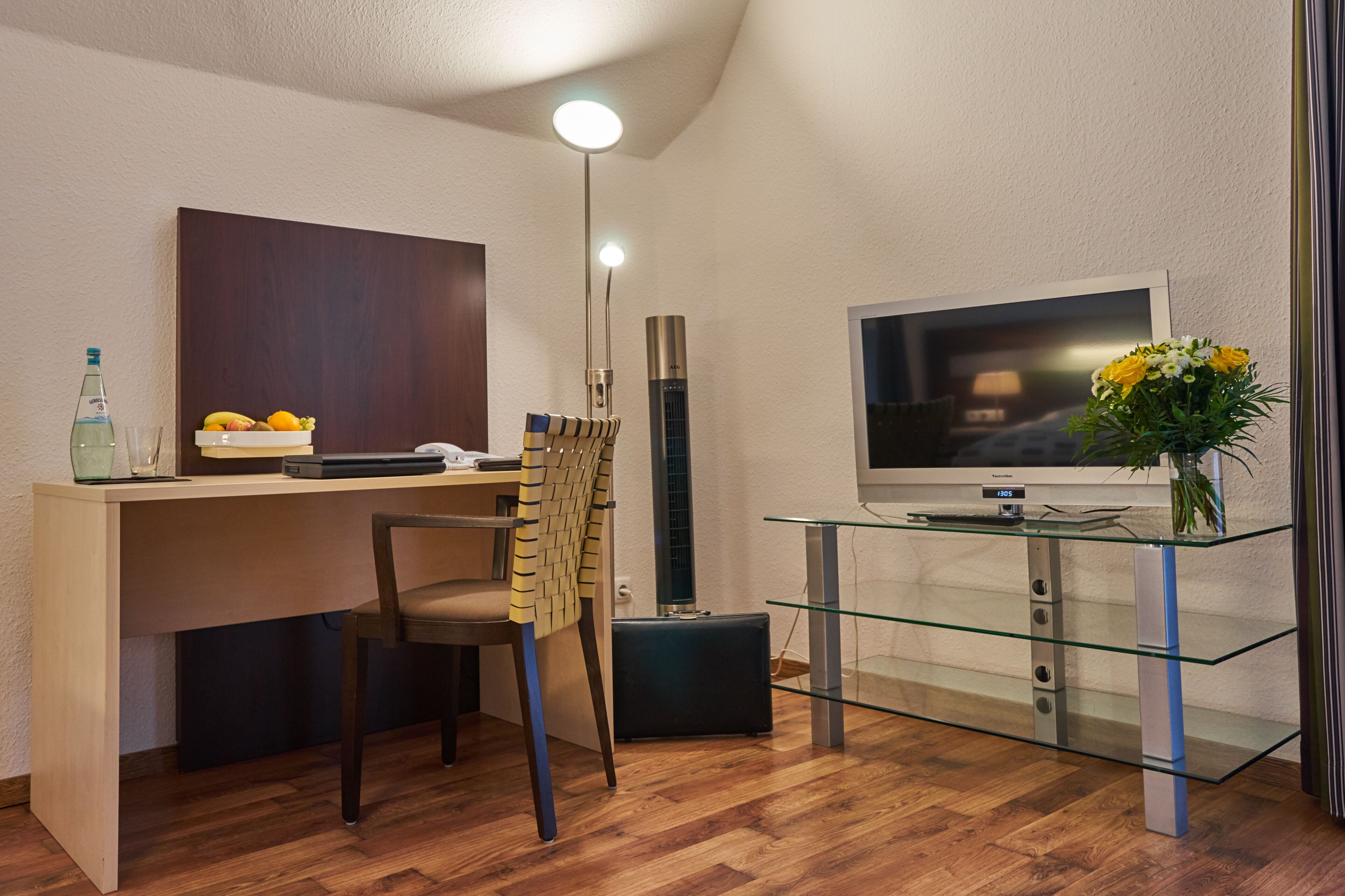 Una sala de estar moderna con un escritorio de madera, una silla, un televisor, una mesa de centro de vidrio y un florero con flores.