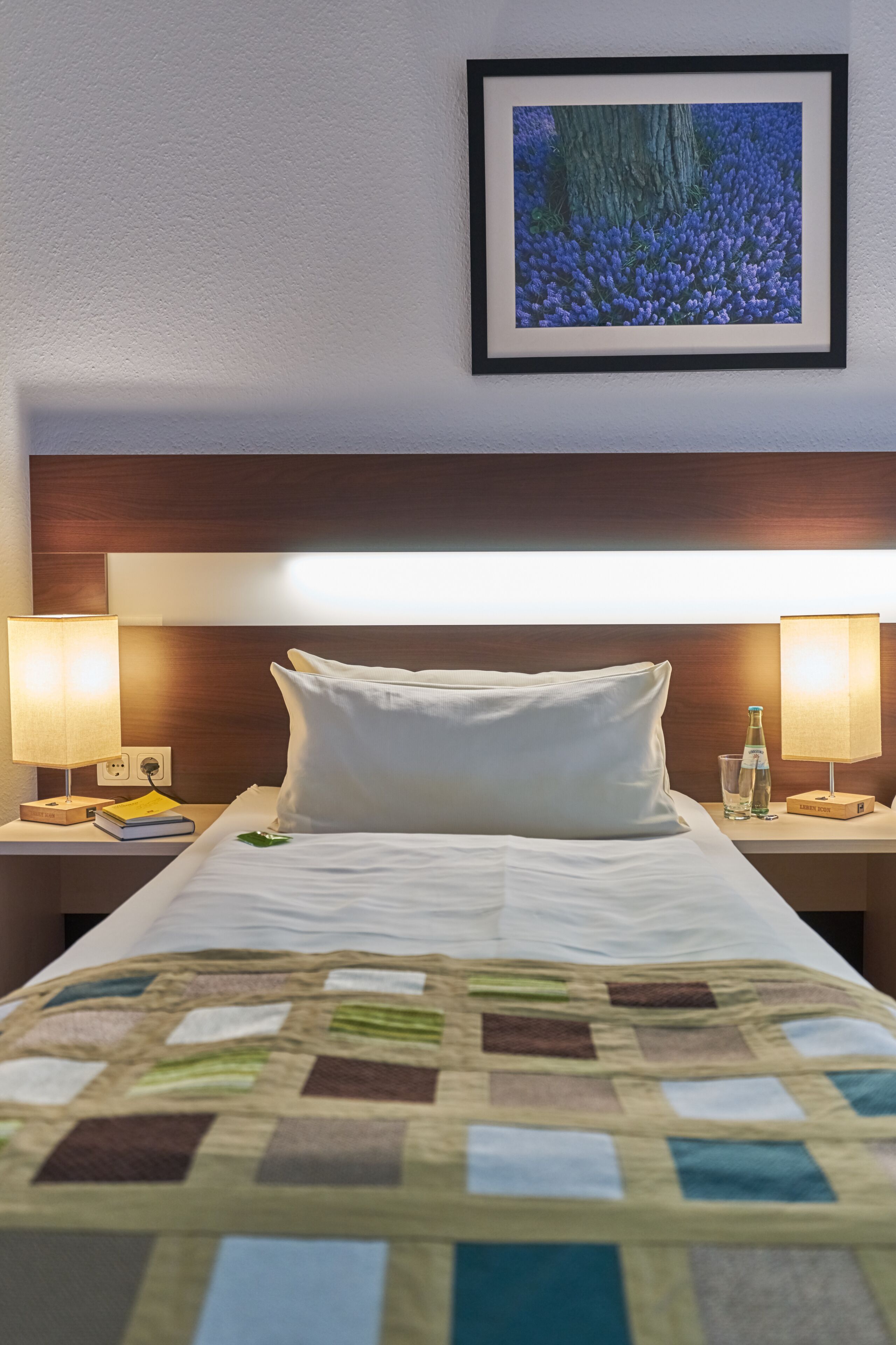 A hotel room with a neatly made bed, two bedside lamps, a framed picture on the wall, and a small table with a book and a bottle.