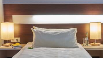 Una habitación de hotel con una cama bien hecha, dos lámparas de noche, un cuadro en la pared y una mesita con un libro y una botella.
