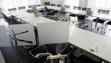 Uma sala de conferências com mesas brancas, cadeiras pretas e um painel de vidro.
