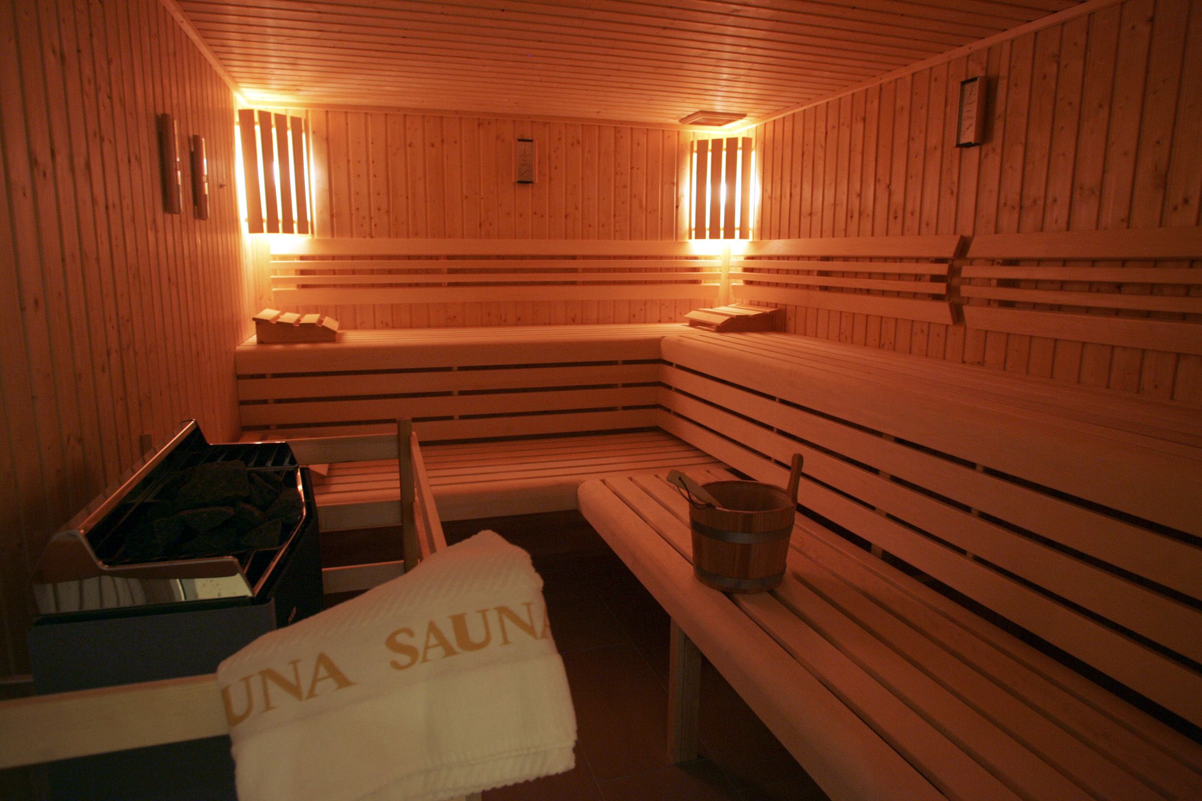 A sauna with wooden benches and benches, a bucket, and a towel with "UNA SAUNA" text.