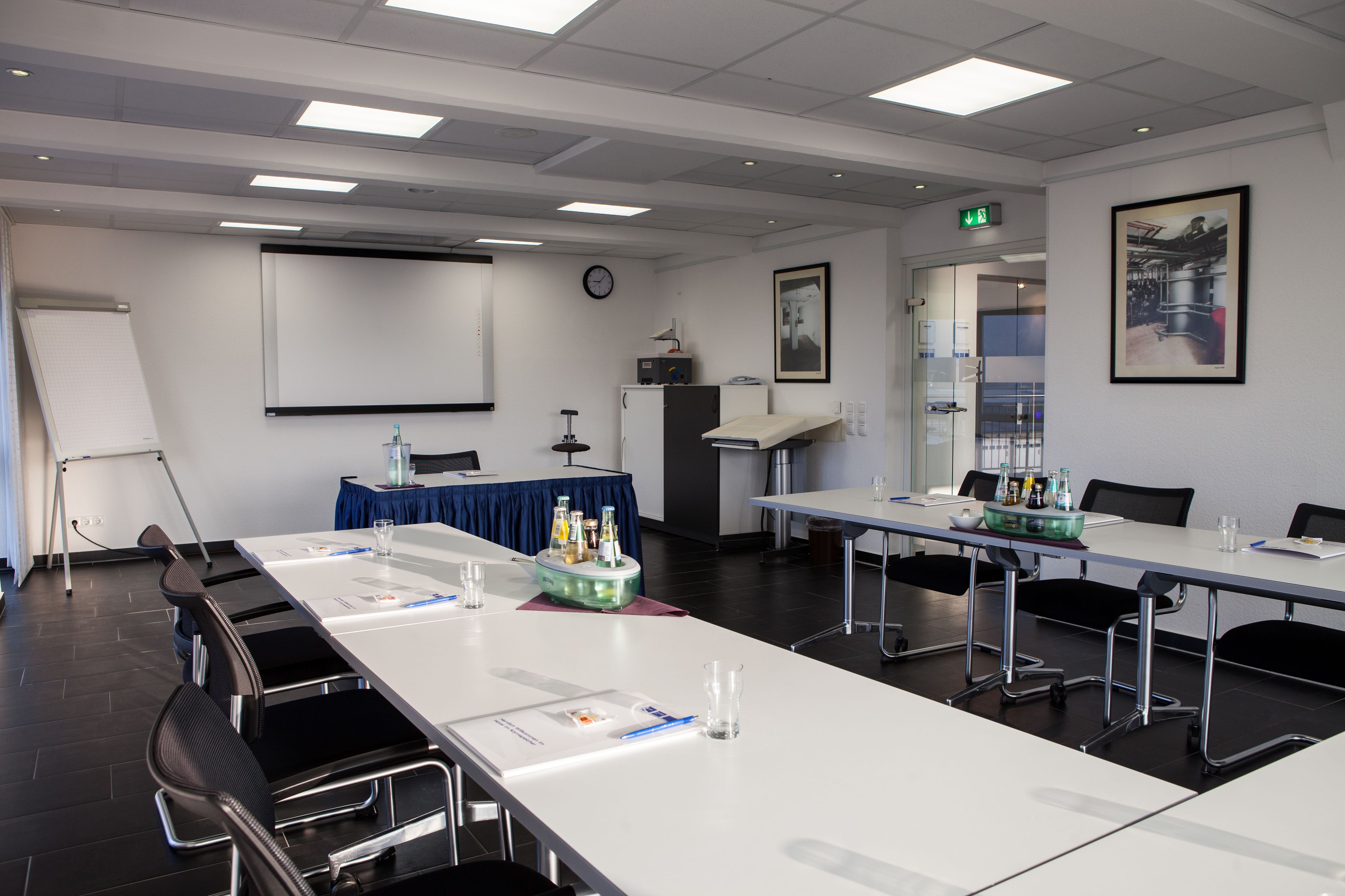 A conference room with a long table, chairs, a projector screen, a clock, and a framed picture on the wall.