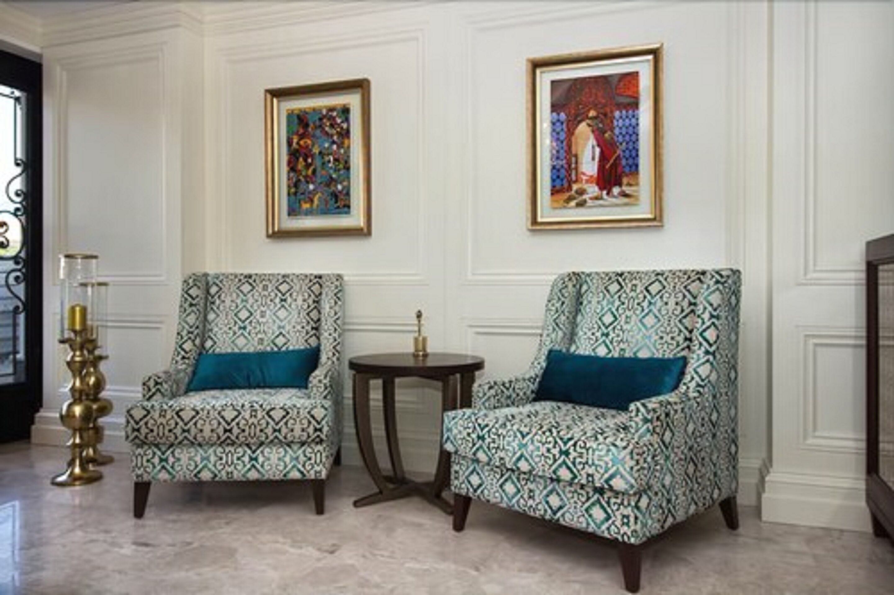 Two patterned armchairs with blue cushions, a small round table, and two framed artworks on the wall.
