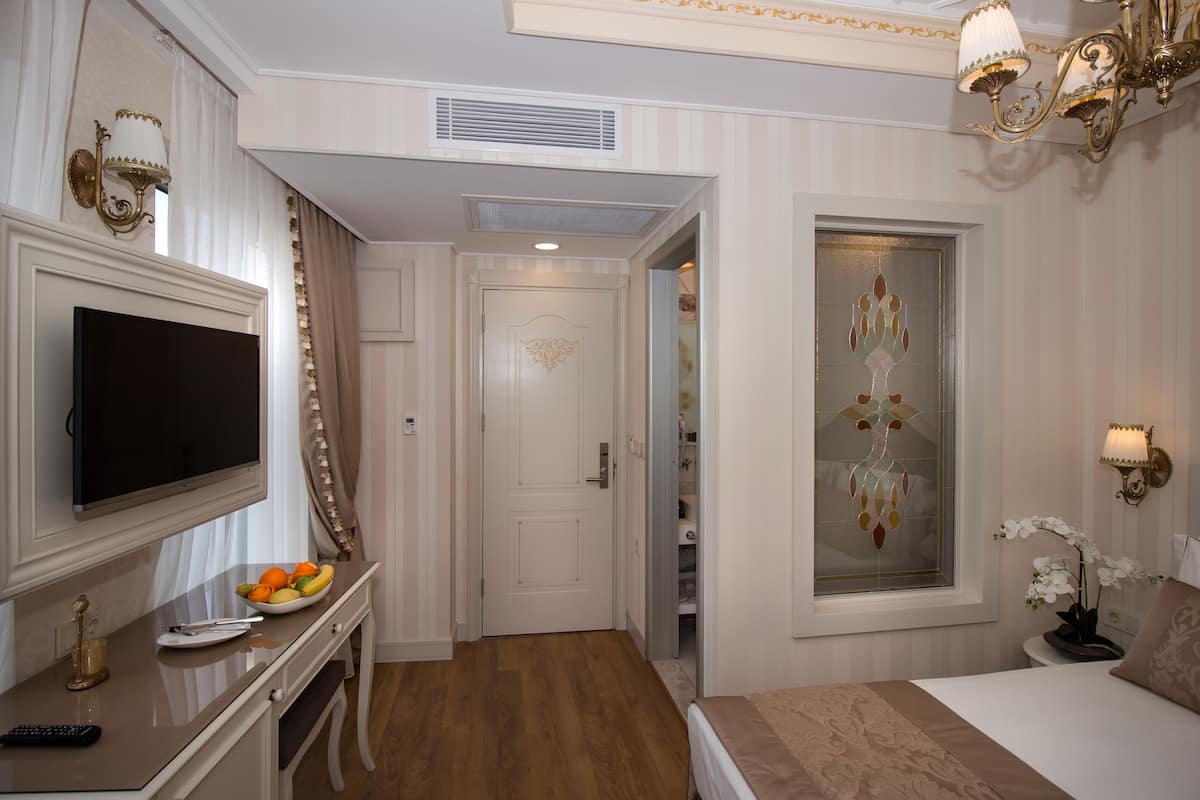 A hotel room with a flat-screen TV, a bedside table with a bowl of fruit, a bed with a decorative headboard, and a window with a decorative glass panel.