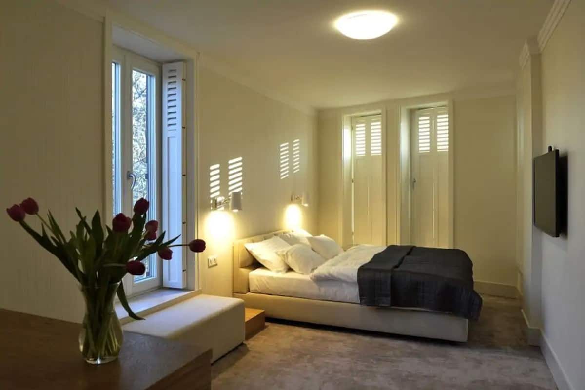 Un dormitorio con cama, mesitas de noche, un jarrón de flores y una ventana con contraventanas.