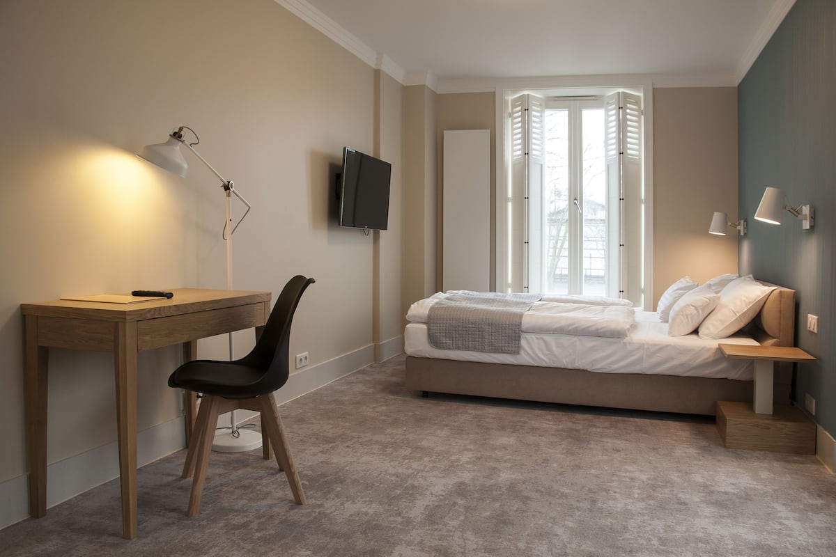 Un dormitorio con cama, escritorio, silla, televisor y ventana con contraventanas.