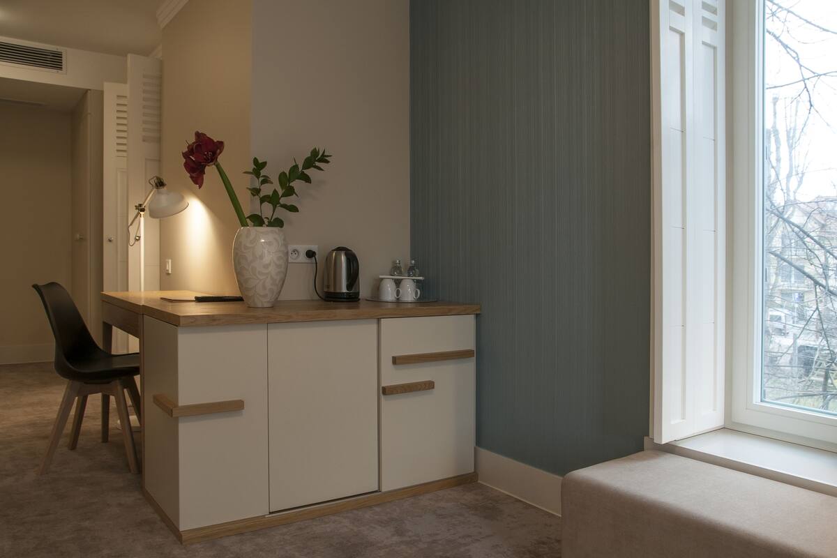Una pequeña kitchenette con encimera de madera, un armario blanco, un jarrón con una rosa roja, una tetera y una cafetera.