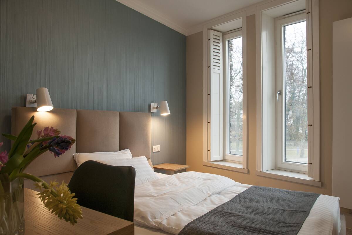 Habitación de hotel con cama, mesitas de noche, una silla y una ventana con contraventanas.