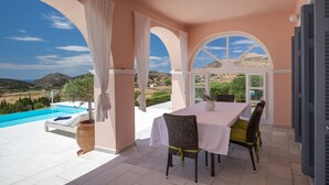 A sunlit patio with a pool, dining table, and chairs, overlooking a scenic landscape.