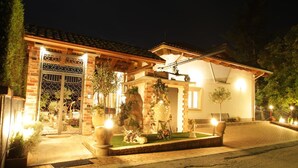 A well-lit residential building with a tiled roof, a gated entrance, and a landscaped front yard with decorative elements. - Wellness Suite Na Canunà (Roddi)