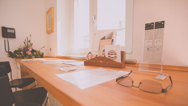 A wooden reception desk with papers, a flower arrangement, and a pair of glasses. - Wellness Suite Na Canunà (Roddi)