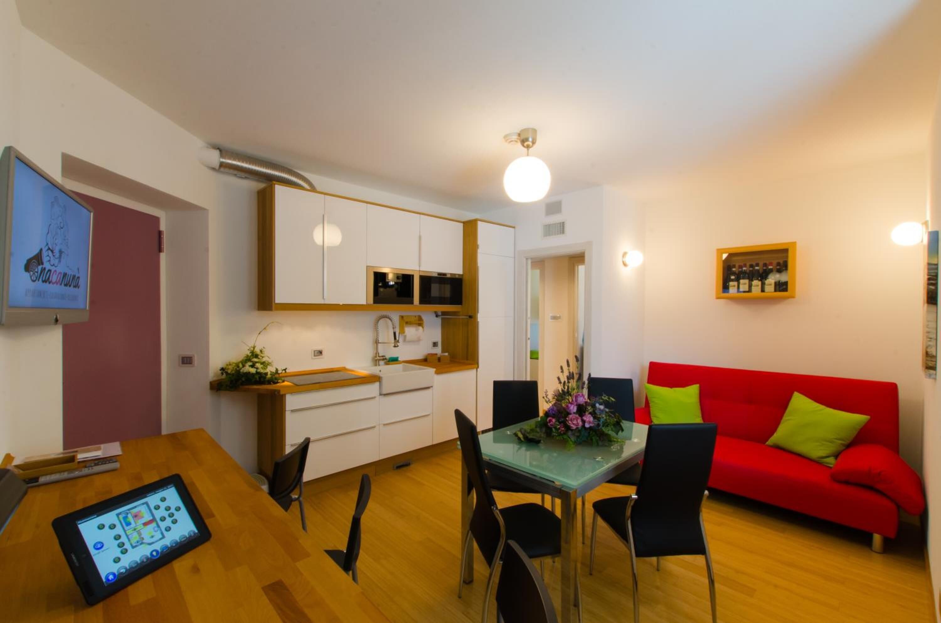 Un salon moderne avec un canapé rouge, une table à manger avec des chaises, un coin cuisine avec des meubles blancs et un téléviseur fixé au mur.