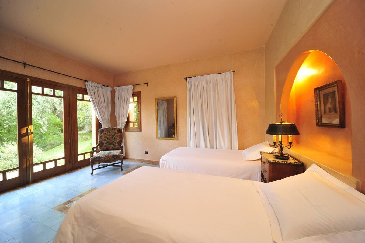 A bedroom with two beds, a chair, a nightstand with a lamp, and a large window with curtains.