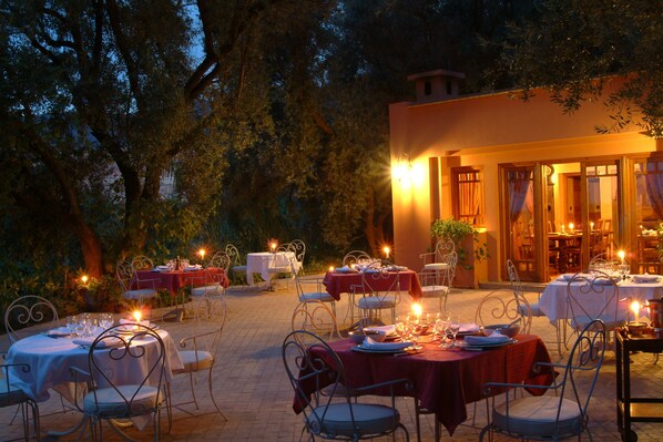 An outdoor dining area with tables set for a meal, surrounded by trees and a building with warm lighting.