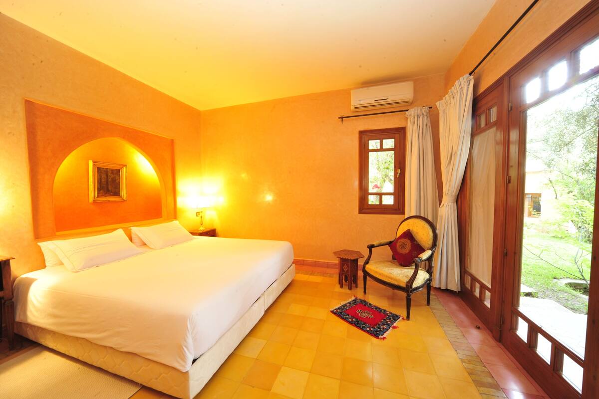A hotel room with a large bed, a chair, a small table, and a window with curtains.
