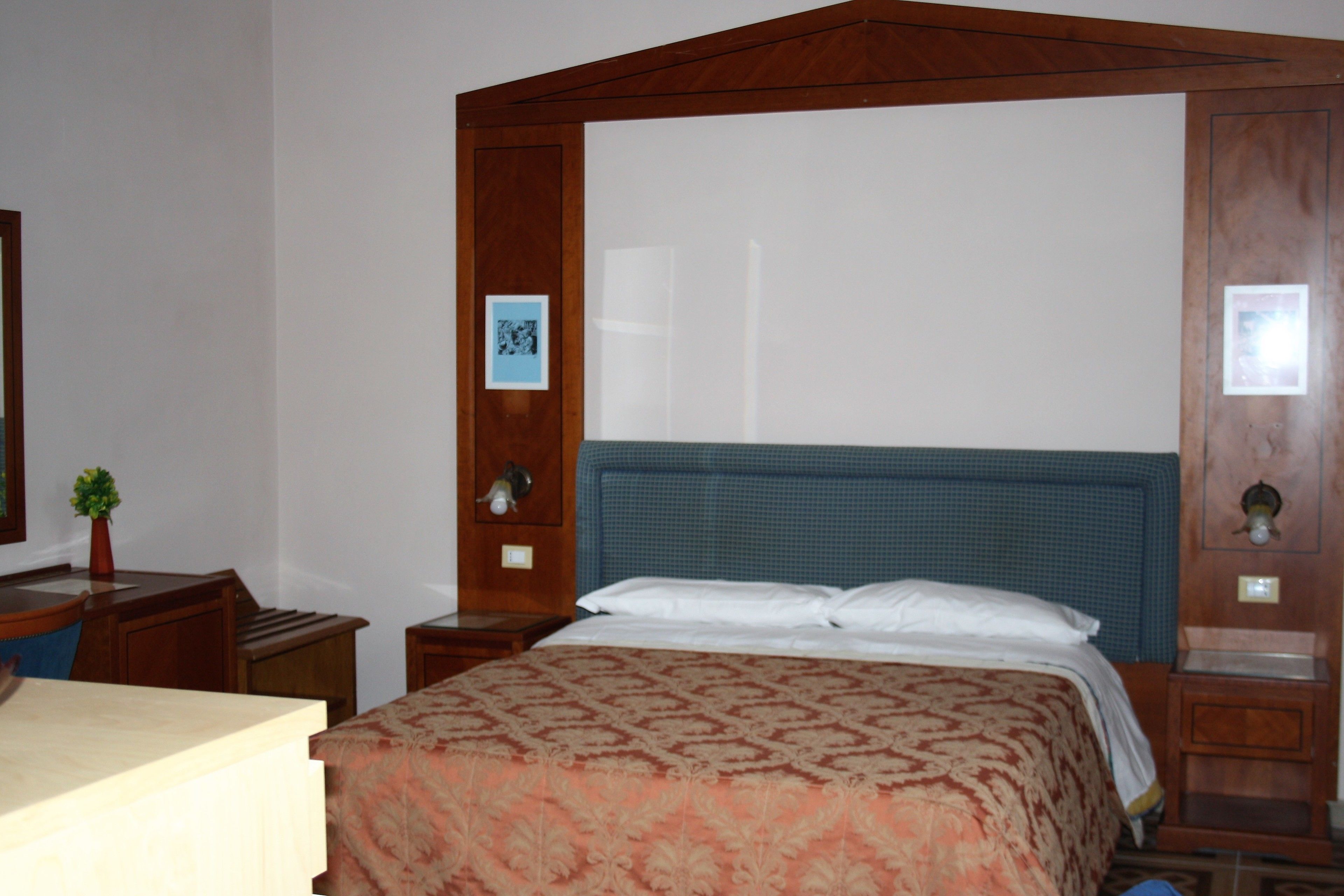 Une chambre à coucher avec une tête de lit en bois, un lit recouvert d’une couvre-lit à motifs, une table de chevet avec une lampe et une petite plante en pot.