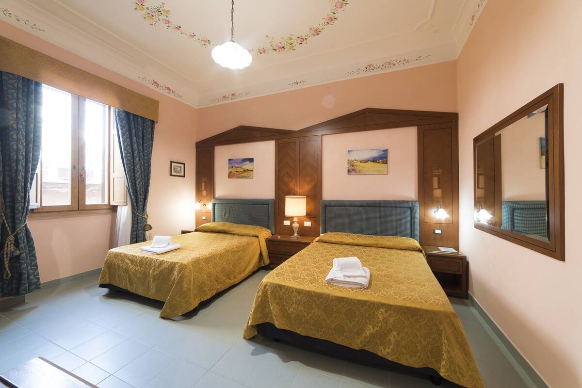 A hotel room with two beds, each with a headboard and a bedside table with a lamp.