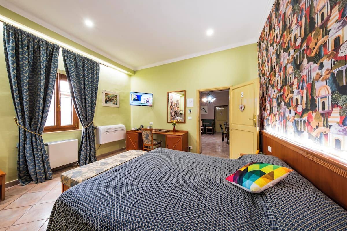 A bedroom with a bed, a desk, a chair, a radiator, a TV, and a wall with a mural.