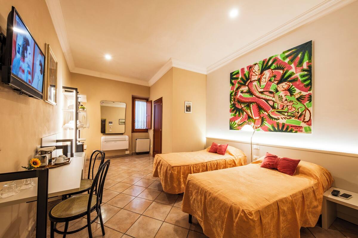 A hotel room with two beds, a dining area with a table and chairs, a TV mounted on the wall, and a large painting on the wall.